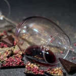 Crveno vino u položenoj čaši sa lomljenom čokoladom posutim sa voćem i začinima oko nje.