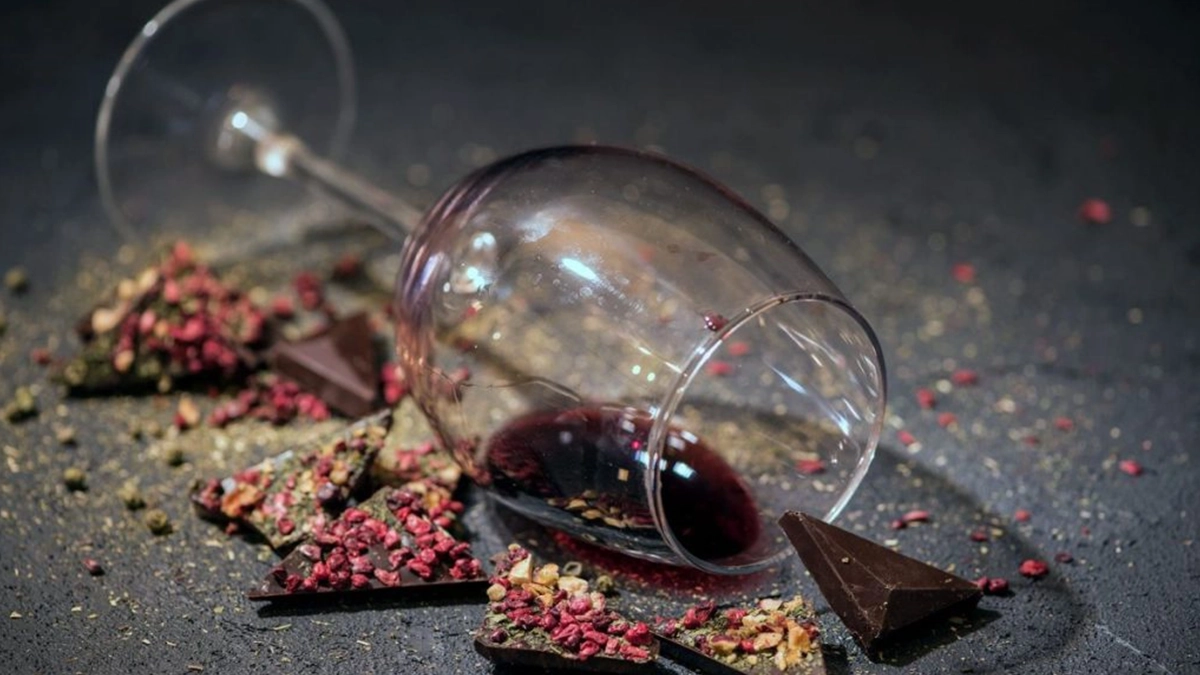 Crveno vino u položenoj čaši sa lomljenom čokoladom posutim sa voćem i začinima oko nje.