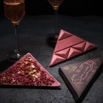 Ruby čokolada sa jagodom, malinom i kajsijom. Pored čokolade na fotografiji se nalaze dve čaše penušavog vina i ambalaža čokolade posvećene vinu Blanc de Noirs.