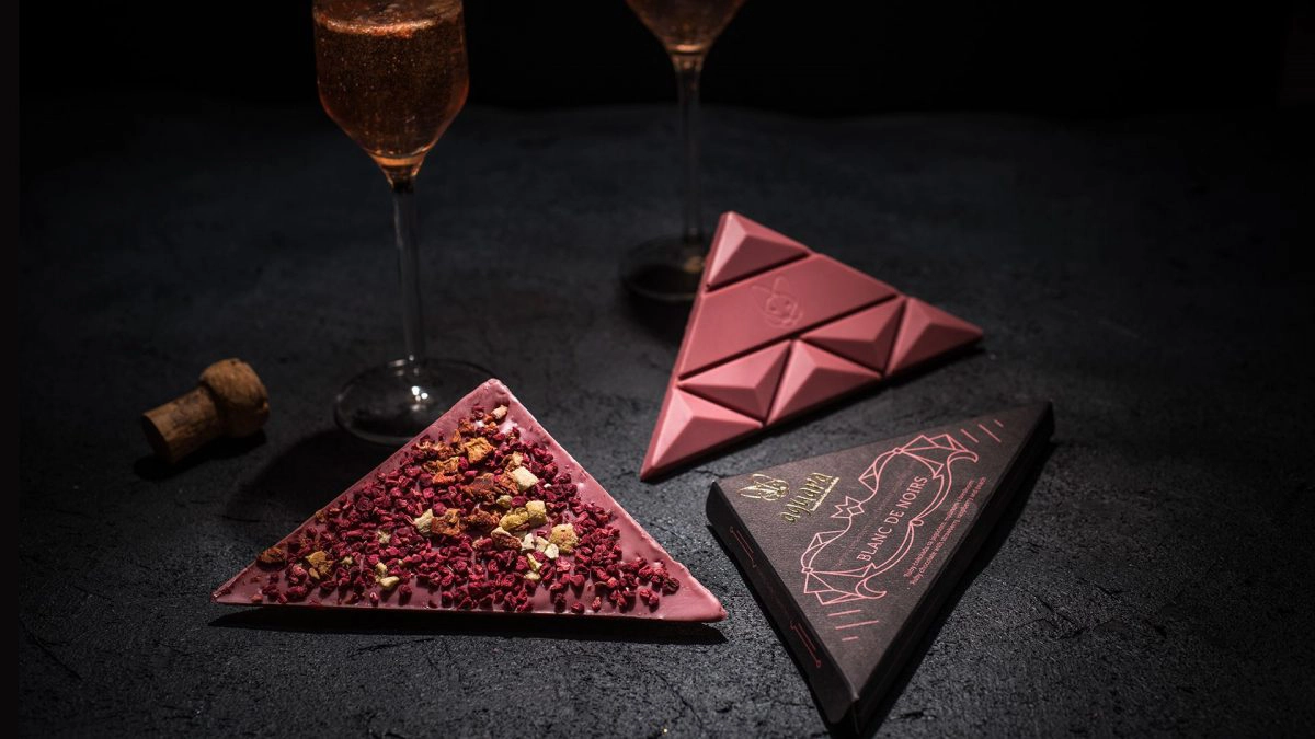 Ruby čokolada sa jagodom, malinom i kajsijom. Pored čokolade na fotografiji se nalaze dve čaše penušavog vina i ambalaža čokolade posvećene vinu Blanc de Noirs.