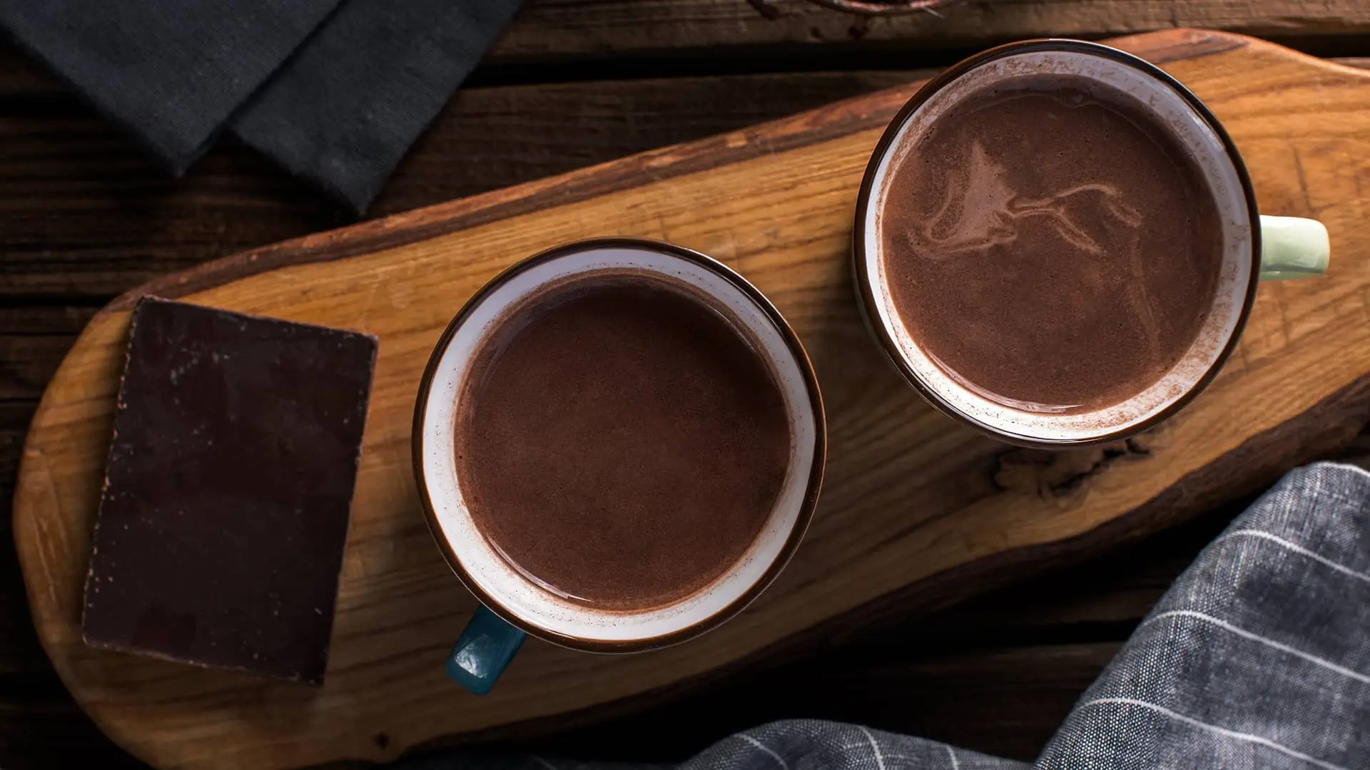 Topla čokolada u dve šolje na drvenoj dasci i komad čokolade pored šolja.